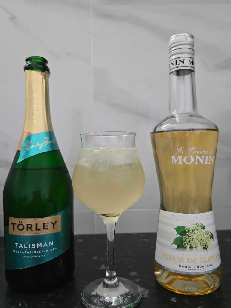 Prikaz hugo koktela i glavnih sastojaka. Sa leve strane se nalazi prosecco, u sredini koktel, a na desnoj strani liker od zove.